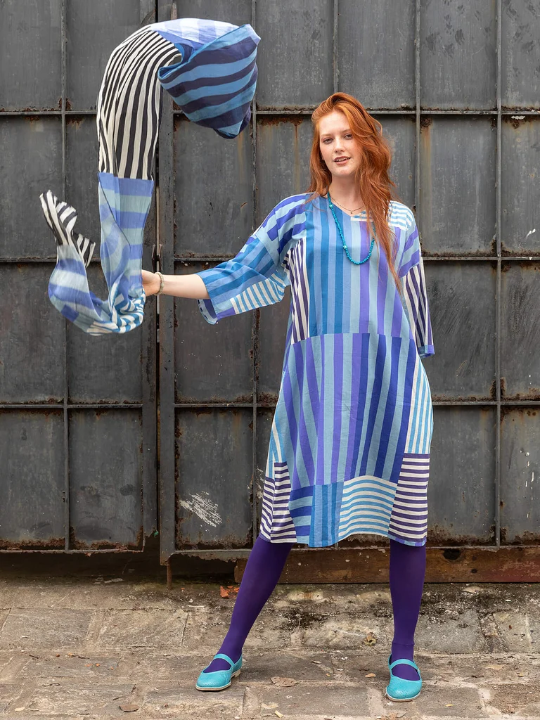 A woman wearing a striped blue dress from Gudrun Sjödén. In her hand, she is holding a matching blue striped shawl from Gudrun Sjödén. 