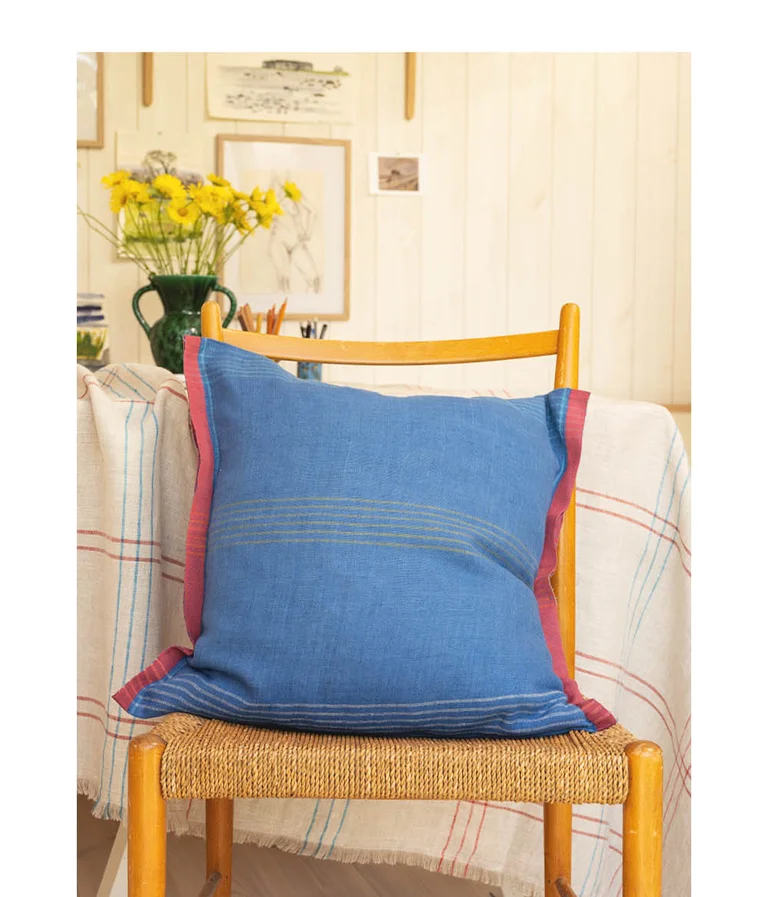 A blue, linen-blend cushion cover with a red selvedge from Gudrun Sjödén on a wooden chair in front of a natural-colour, check linen textile.