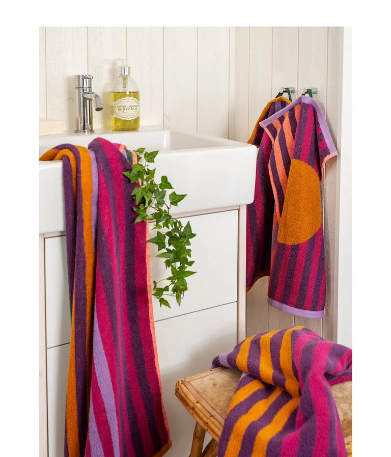 Purple, cerise and orange striped terry towels from Gudrun Sjödén, hooked next to a washbasin and draped over a stool. One of the guest towels has a large orange dot.