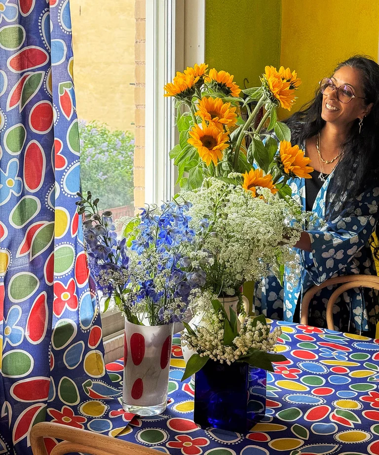 A woman in a patterned blue and white kimono from Gudrun Sjödén arranging flowers on a table with a colorful patterned tablecloth from Gudrun Sjödén.