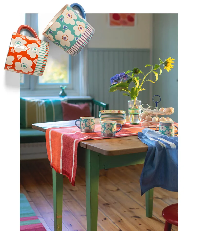 A kitchen table laid with red and blue striped textiles from Gudrun Sjödén, floral mugs and a bunch of flowers. Floral ceramic mugs from Gudrun Sjödén are superimposed on the image.