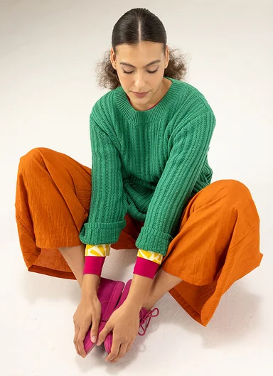 A seated woman with a green knit sweater and orange pants from Gudrun Sjödén.