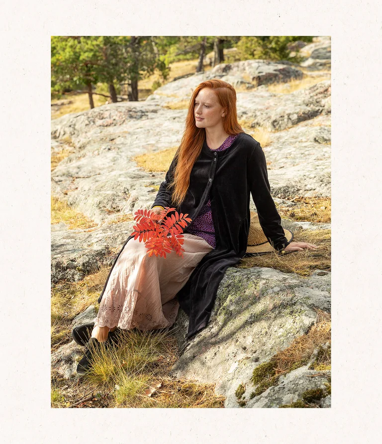 A woman sitting down and wearing a buttoned black velour dress from Gudrun Sjödén. She is holding a branch from a mountain ash tree in her hand.