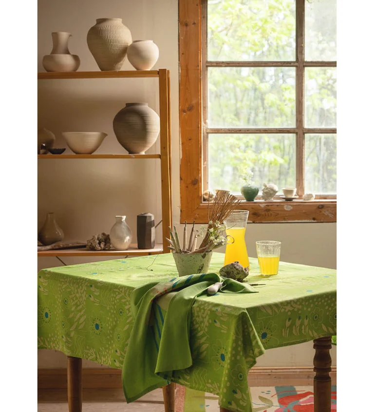 A table with a pea green cloth from Gudrun Sjödén in a pottery studio, in front of shelves filled with ceramic pots and a Georgian window overlooking greenery.