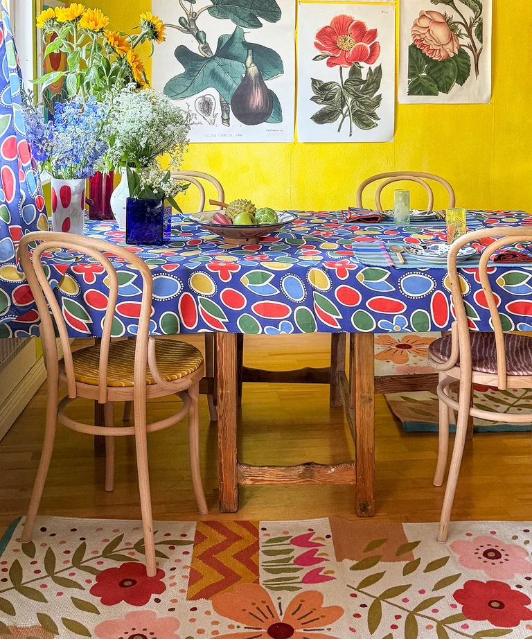 Dining table with a patterned tablecloth in cobalt blue from Gudrun Sjödén. There are botanical posters on the yellow wall, and there is a floral rug from Gudrun Sjödén under the table.