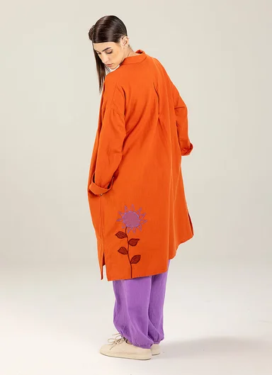A woman wearing an orange coat with an embroidered pattern and a pair of purple pants from Gudrun Sjödén.