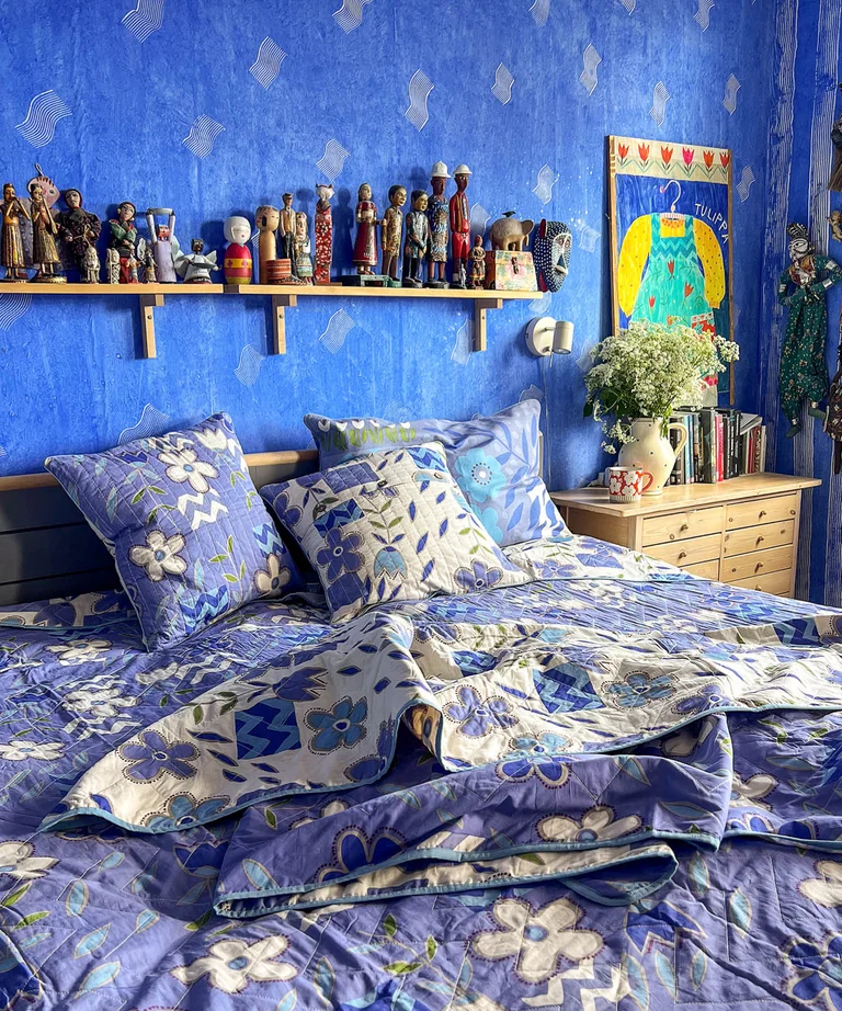 Double bed with a bedspread and cushion covers in blue and purple from Gudrun Sjödén, in front of an ultramarine-blue wall with a small shelf filled with dolls and figurines.