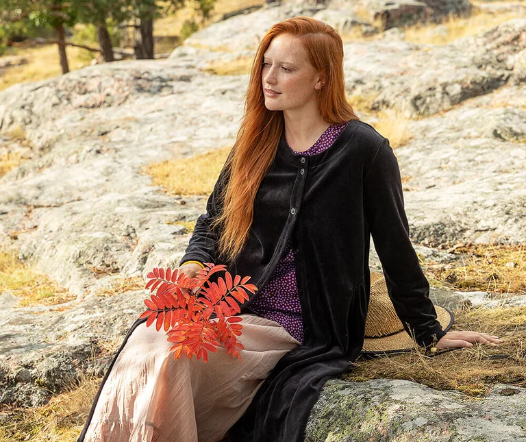 A woman sitting on a rock wearing a black velour dress and a patterned top from Gudrun Sjödén, holding a twig of rowan berries.