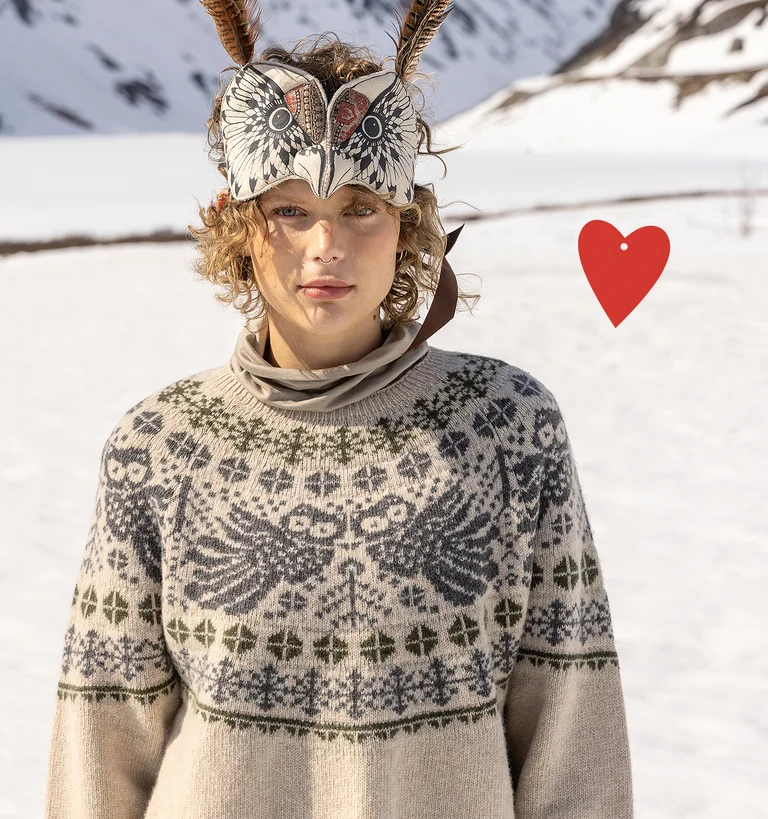 A woman with an animal mask on her head standing in a snowy landscape wearing layers of clothing from Gudrun Sjödén’s holiday collection 2025.  